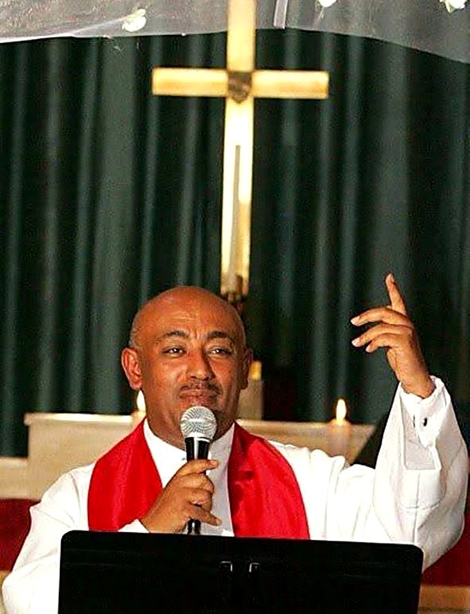 Rev. Dr. Dereje Fantaye Yimer preaching beneath the cross, a red stole over his white robe, the microphone in one hand and the other raised.